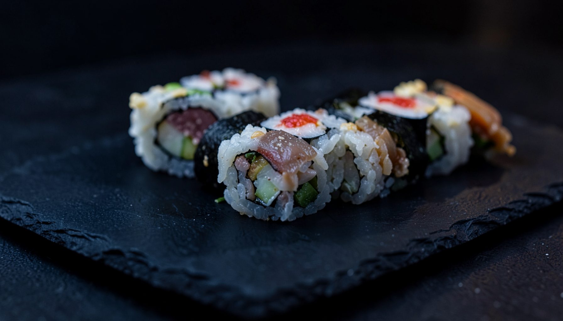 Assorted sushi rolls arranged on a dark slate plate with deep navy and charcoal tones